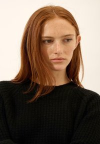 Czarny sweter z fakturowanym prążkowanym wzorem, z okrągłym dekoltem. Modelka ma długie czerwone włosy i widoczne piegi na twarzy.
