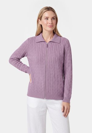 Vrouw draagt een lichtpaarse kabelgebreide trui met rits en kraag en witte broek, poseert met één hand op de heup tegen een witte achtergrond.