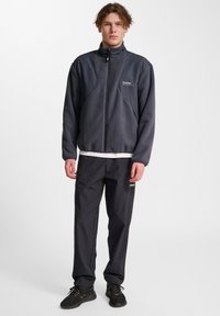 Fleecetröja i mörkgrått med dragkedja fram, hög krage och logotyp på bröstet, kombinerad med matchande mörka byxor och svarta sportskor.