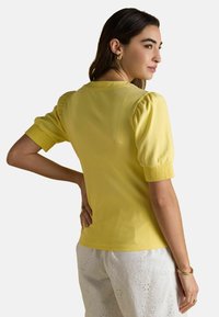 Naisel, kellel on pikad tumedad juuksed, on seljas kollane puffvarrukatega topp ja valged silmadega püksid, seistes käsi puusal, vaadates külje poole.