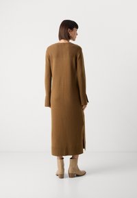 Vestito lungo marrone in maglia con texture a coste nella parte superiore e nelle maniche, dotato di spacchi laterali. Il modello indossa stivaletti beige alla caviglia.