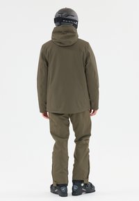 Olivgrüne Skijacke mit Kapuze, kombiniert mit passenden Hosen. Aus wasserdichtem Material und schwarzen Skistiefeln mit verstellbaren Schnallen.