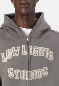 Siva zip-up dukserica s teksturiranim natpisom "LOW LIGHTS STUDIOS" s metalnim akcentima. Mekani materijal s opuštenim krojem i velikom kapuljačom.