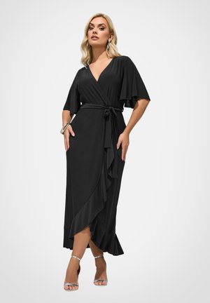 Lyshåret kvinde i sort omslagskjole med flæsekant og flagrende ærmer, accessoriseret med sølvarmbånd, øreringe og åbne hæle.