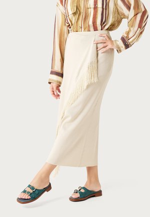 Vrouw draagt een beige wikkelrok met franjes, een gestreept overhemd met knopen en teal sandalen met open teen en gouden gespen, staand met één hand op de heup.