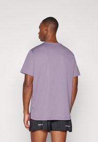 Lilla t-shirt med korte ærmer og rund hals, glat tekstur og sideslids, parret med sorte shorts med et diskret mønster.
