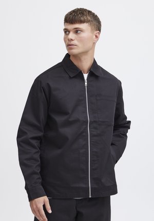 Ung mand iført en sort lynlåsjakke med brystlomme, kigger mod venstre mod en ensfarvet hvid baggrund.