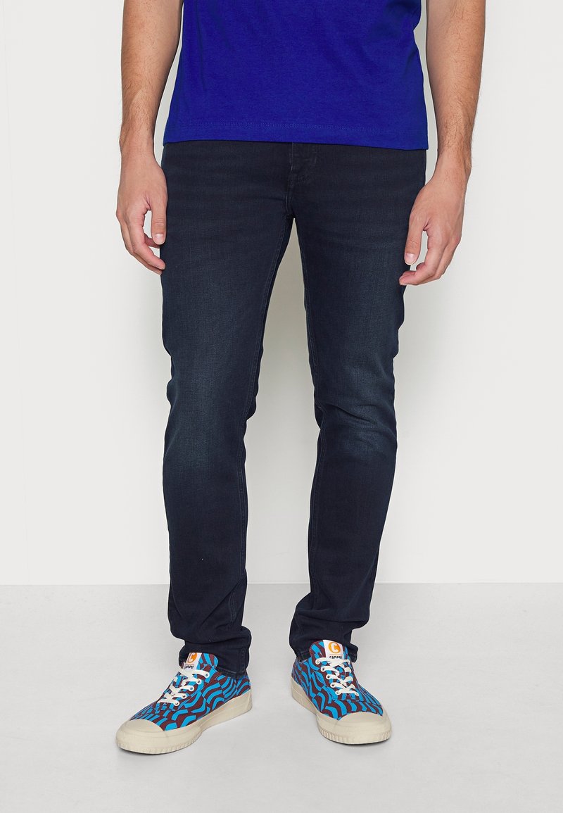 Vaqueros azul oscuro de corte slim, combinados con zapatillas azules que presentan un patrón ondulado y suelas blancas. La camisa es de un azul real sólido.