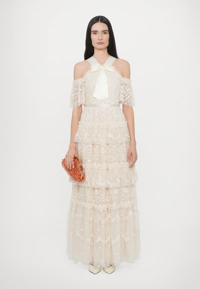 Crèmekleurige maxi-jurk van kant met off-shoulder mouwen, tiered ontwerp gedetailleerd met bloemenpatronen, gecombineerd met een koraalkleurige beaded clutch en beige hakken.
