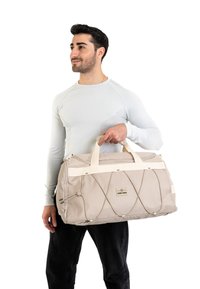 Borsa duffel beige con manici bianchi, superficie texturizzata e dettagli in rete elastica. La borsa presenta un'etichetta logo sul davanti.