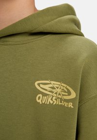 Olívazöld kapucnis felső, bézs elülső logóval, puha tapintású, letisztult dizájnnal és kenguruzsebbel.