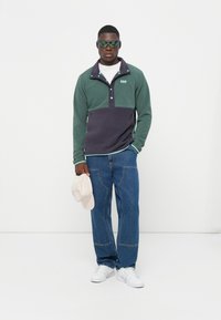 Pullover in donker groen en marineblauw, met een witte kraag en knopenlijst. Draagt bij blauwe jeans en witte sneakers. Accessoire: lichtbeige pet.