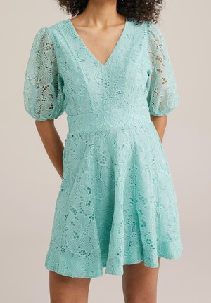 Kvinde iført en mintgrøn kjole med blonder, puffede ærmer til albuen og V-udskæring, stående foran en beige baggrund.