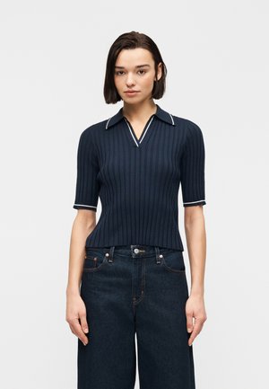 Jeune femme aux cheveux raides et foncés porte un polo côtelé bleu marine à manches courtes et un jean large taille haute foncé, debout devant un fond blanc.