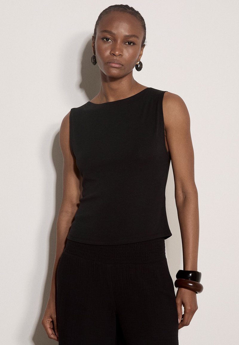 Femme aux cheveux tressés porte un haut noir sans manches, un pantalon noir, des boucles d'oreilles en anneau et de larges bracelets noirs et marron au poignet droit.