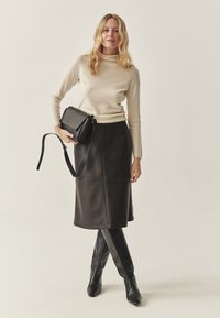 Fekete bőr szoknya, bézs testhez simuló pulóver és fekete térdig érő csizma. A modell egy kis fekete válltáskát tart pánttal. Texturált, minimalista dizájn.