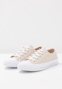 Zapatillas de deporte con cordones y una parte superior de color beige texturizada, puntera de goma blanca y suela. Presenta ojales plateados y cordones blancos planos.