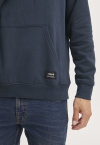 Sudadera azul marino con un bolsillo frontal, puños y dobladillo acanalados. Presenta una etiqueta de la marca visible en la parte inferior izquierda. Textura suave.