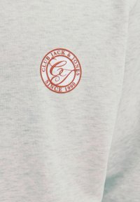 Vaaleanharmaa puuvillakangas, jossa on hienovarainen tekstuuri ja keskellä pyöreä punainen logo, jossa lukee "CLUB JACK & JONES SINCE 1990".