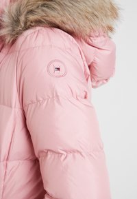 Różowa kurtka puchowa z futrzanym kapturem, pikowaną fakturą i haftowanym logo na lewym rękawie.