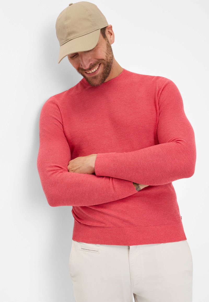 Roter gestrickter Pullover mit rundem Kragen, tailliertem Schnitt und gerippten Bündchen. Kombiniert mit hellen Hosen und einer beigen Kappe.