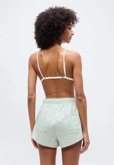 Γυναίκα με σγουρά μαλλιά που φοράει λευκό σουτιέν και κοντό παντελόνι με ελαστική μέση σε ανοιχτό πράσινο χρώμα, εμφανιζόμενη από πίσω σε ουδέτερο φόντο.