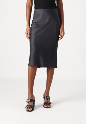 Kobieta ubrana w czarną spódnicę midi i czarne sandały na obcasie ze złotymi klamrami, stojąca na jednolitej białej podłodze i tle.