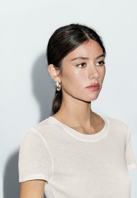 Jonge vrouw met donker haar in een staart, die zilveren oorringen draagt en een witte, getailleerde top met korte mouwen aan heeft, kijkt naar rechts tegen een lichte achtergrond.