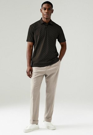 Schwarzes Poloshirt mit kurzen Ärmeln und geometrischem Muster, kombiniert mit hellbeigen Hosen und weißen Turnschuhen. Glatter Stoff und klassische Passform.