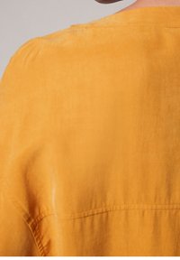 Blusa gialla caratterizzata da una texture morbida e liscia. Il tessuto si drappeggia in modo ampio con una vestibilità rilassata e include un dettaglio sottile della cucitura sulla spalla.