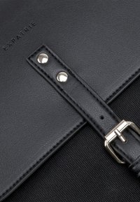 Svart läderväska med en texturerad yta, utrustad med ett band med silverfärgad hårdvara, nitar och präglad "EXPATRIÉ"-logotyp.