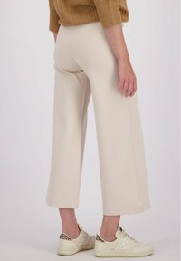 Vrouw in beige wijdvallende cropped broek, een bruine trui en lichtgekleurde sneakers met leopardenprintdetails, staand tegen een witte achtergrond.