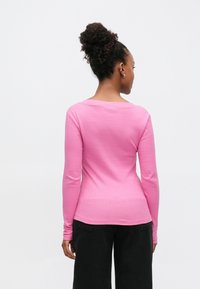 Langärmliges, geripptes T-Shirt in leuchtendem Pink. Mit weitem Ausschnitt und figurbetonter Silhouette. Kombiniert mit schwarzen, weit geschnittenen Hosen.