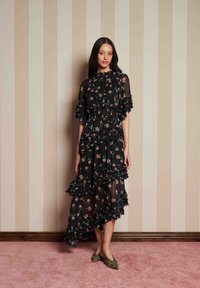 Kvinna i en genomskinlig svart blommig klänning med volanger och leopardmönstrade spetsiga skor som står mot randig beige tapet.