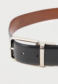 Črni usnjen pas z gladko teksturo, z srebrno kovinsko sponko. Notranja podloga je rjava in ima več lukenj za nastavitev velikosti.