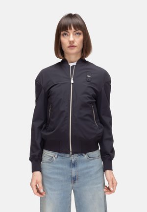 Donna con capelli castani lisci indossa una giacca bomber nera con cerniera e jeans azzurri, in piedi davanti a uno sfondo bianco semplice.