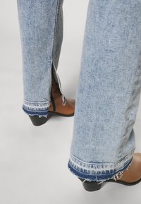 Jean en denim bleu clair avec ourlets effilochés et fentes latérales porté avec des bottines marron à talons sur un sol gris.