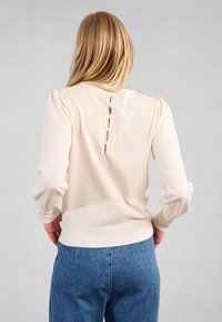 Maglione lavorato a maglia color crema con maniche a sbuffo e chiusura posteriore con quattro anelli di tessuto e bottoni. Design strutturato e aderente.