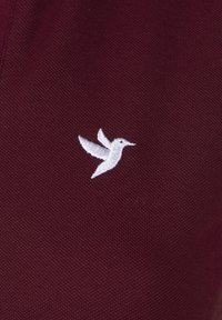 Burgunder stof med en struktureret vævning, der har et hvidt broderet kolibri-logo, placeret fremtrædende på overfladen.