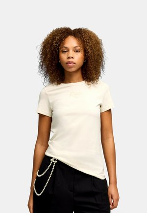 Mujer con cabello rizado que lleva una camiseta Puma beige claro y pantalones negros con un accesorio decorativo de cadena de perlas.