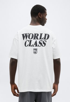 Man die wegkijkt en een wit T-shirt draagt met grote zwarte tekst "WORLD CLASS" en een klein embleem eronder tegen een effen achtergrond.