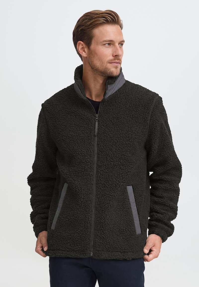 Homme portant une veste polaire noire zippée avec un col montant et des accents gris sur les poches, regardant sur le côté sur un fond uni.