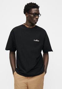 Άνδρας που φοράει μαύρο T-shirt Carhartt και μπεζ παντελόνι, κοιτάζοντας προς τα κάτω και στο πλάι, με τα χέρια στις τσέπες, σε απλό φωτεινό φόντο.