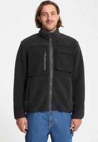 Veste en polaire noire avec un panneau en tissu lisse sur le haut de la poitrine, poches zippées et col montant. Elle est chaude et texturée.