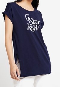Námořnické modré tričko vyrobené z bavlny, s kulatým výstřihem a ohrnutými krátkými rukávy. Velké bílé logo "G-Star RAW" tištěné na přední straně.