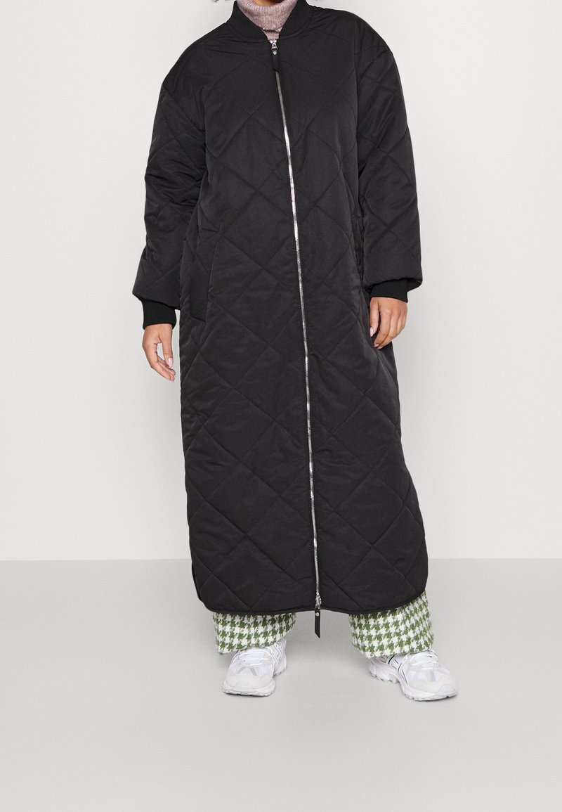 Henkilö, jolla on päällään pitkä musta päällystakki, jossa on vetoketju edessä, vihreät ja valkoiset kuviolliset housut, valkoiset lenkkarit ja vaaleanpunainen poolopaita.