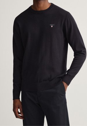Homme portant un pull noir uni à col rond avec un petit logo GANT brodé sur la poitrine et un pantalon noir, main dans la poche.