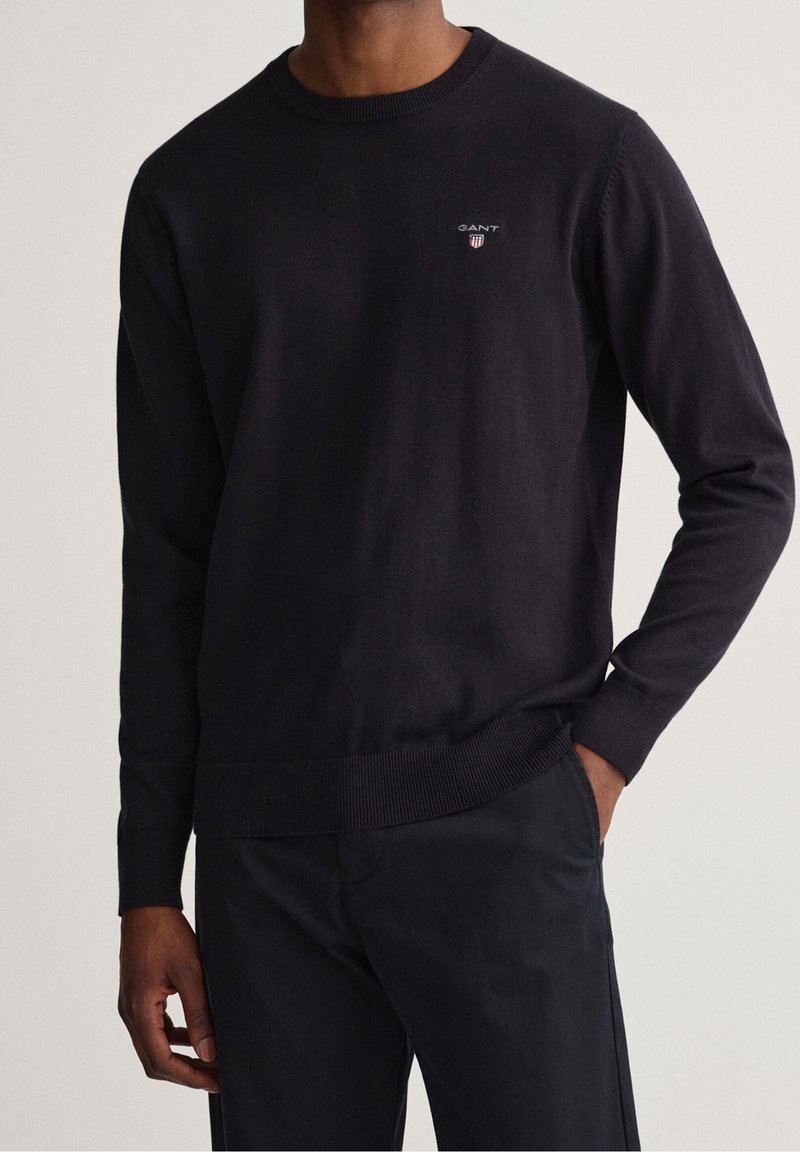 Homme portant un pull noir uni à col rond avec un petit logo GANT brodé sur la poitrine et un pantalon noir, main dans la poche.