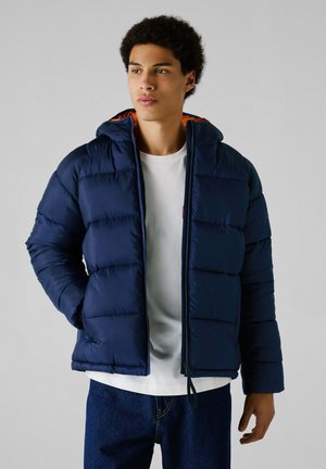 Marineblaue Steppjacke mit gefütterter Kapuze, gestepptem Design und Reißverschluss; getragen über einem weißen Hemd und kombiniert mit dunklen Jeans.