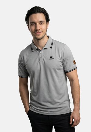 Mann mit dunklem Haar, der ein hellgraues Forsberg-Poloshirt mit schwarzem Besatz und Logo auf Brust und Ärmel trägt, steht mit einer Hand in der Tasche.